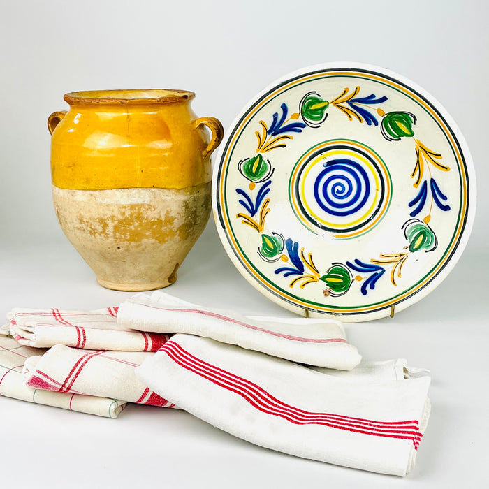 Spanish folk art lebrillo paired with a yellow earthenware vase and colorful striped cloths, highlighting the hand painted bowl as an early 20th century decorative object. Its central blue spiral, green floral wreath and concentric bands create a strong visual anchor for Mediterranean decor, a ceramic collection or a kitchen wall display.