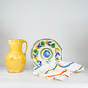 Spanish folk art lebrillo paired with a yellow earthenware vase and colorful striped cloths, highlighting the hand painted bowl as an early 20th century decorative object. Its central blue spiral, green floral wreath and concentric bands create a strong visual anchor for Mediterranean decor, a ceramic collection or a kitchen wall display.