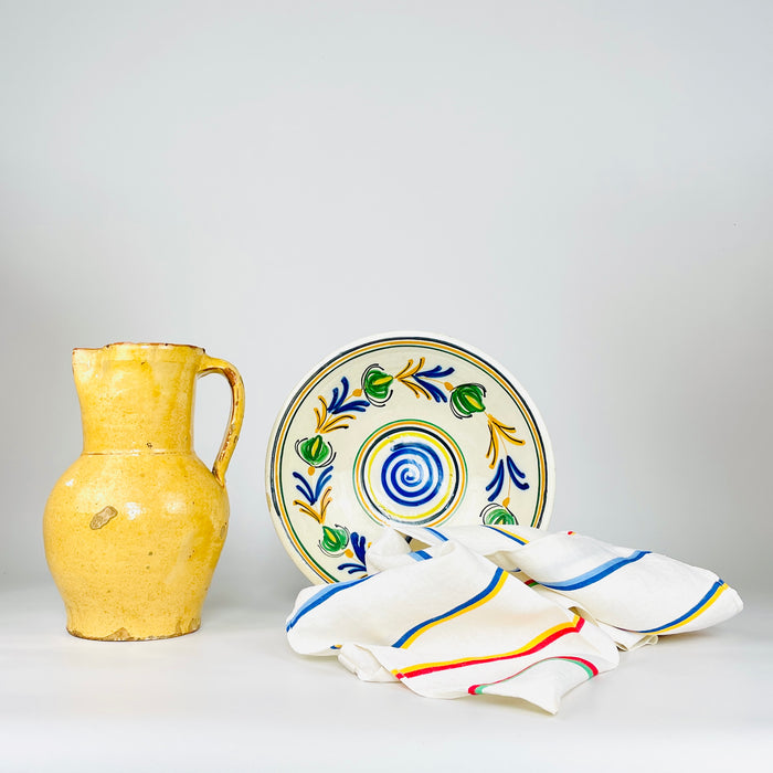 Spanish folk art lebrillo paired with a yellow earthenware vase and colorful striped cloths, highlighting the hand painted bowl as an early 20th century decorative object. Its central blue spiral, green floral wreath and concentric bands create a strong visual anchor for Mediterranean decor, a ceramic collection or a kitchen wall display.