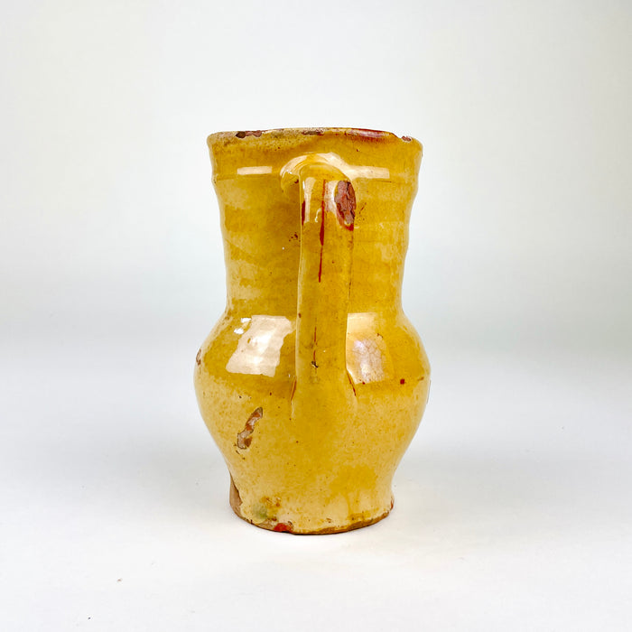 Antique Castilian pottery pitcher with a hand-shaped pouring spout and applied handle joining the shoulder to the rim. The red terracotta body is covered in a translucent honey yellow glaze that pools around the neck and handle, with extensive age-related rubbing, chips and surface crazing visible across this Spanish folk art vessel.