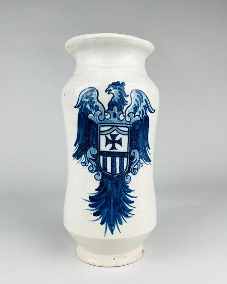 Antique Talavera style Spanish apothecary jar with a centrally placed heraldic eagle painted in deep and light cobalt blue on a creamy white ceramic ground. The eagle has extended wings, ornamental scrolls and a long feathered tail surrounding its shield. This substantial albarello vessel combines a traditional pharmacy jar silhouette with graphic historic decorative motifs.