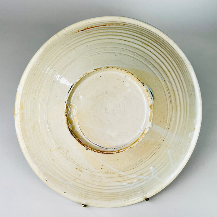 Reverse of an antique Spanish lebrillo bowl showing the cream glazed earthenware body, circular foot ring and pronounced concentric throwing ridges left by wheel construction. Irregular glaze pooling, kiln traces and age related surface wear document handmade manufacture, while the broad base confirms the piece was formed as a functional shallow basin.