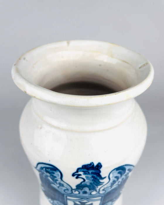 Hand painted cobalt blue heraldic eagle on a vintage Spanish Talavera albarello jar, showing its hooked beak, feathered head and raised wings. Several blue tones define the bird against the white glazed earthenware, with darker outlines and lighter interior brushwork. The eagle rests above a shield containing a cross, giving this decorative pharmacy vessel a striking heraldic identity.