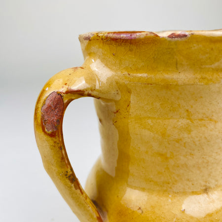 Spanish terracotta jug sourced in Peñafiel, Valladolid, measuring 15.5 cm high and 12 cm wide from spout to handle. Its wheel-thrown earthenware form carries a mottled yellow glaze over red clay, with a bulbous lower body, narrow waist and practical pouring lip characteristic of traditional Castilian folk pottery.