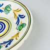 Antique Spanish remolino lebrillo displayed with a yellow glazed confit pot and striped linen textiles, demonstrating the bowl’s scale and vivid color relationship. The 28.5 centimeter ceramic piece combines cobalt blue, green, ochre and black over cream glaze, making it a practical choice for farmhouse decor, open shelving or a collected pottery arrangement.