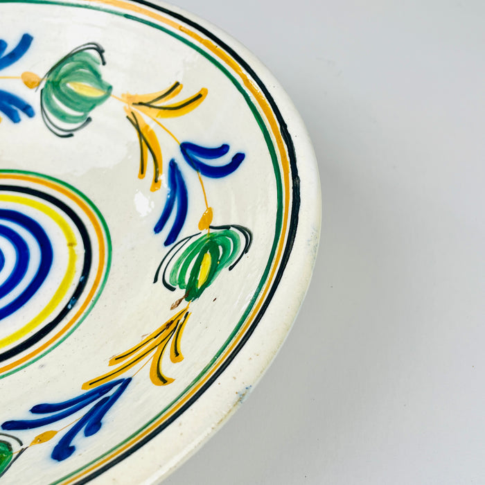 Antique Spanish remolino lebrillo displayed with a yellow glazed confit pot and striped linen textiles, demonstrating the bowl’s scale and vivid color relationship. The 28.5 centimeter ceramic piece combines cobalt blue, green, ochre and black over cream glaze, making it a practical choice for farmhouse decor, open shelving or a collected pottery arrangement.