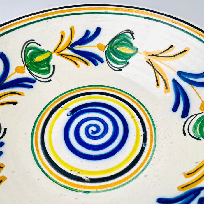 Antique Castilian remolino bowl in hand painted glazed earthenware, its broad interior organized into circular decorative zones. A vivid cobalt spiral sits inside yellow, black, ochre and green rings, surrounded by repeating blue leaves and green pod shaped flowers. The form serves equally well as a decorative bowl or plate wall centerpiece.