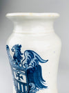 Lower portion of the heraldic decoration on a Spanish blue and white albarello apothecary jar, featuring a shield with a dark cobalt cross and vertical stripes. Curled scroll ornaments frame the shield, while long layered eagle tail feathers extend down the white ceramic body. The hand painted design provides an elaborate graphic contrast to the simple wheel thrown form.