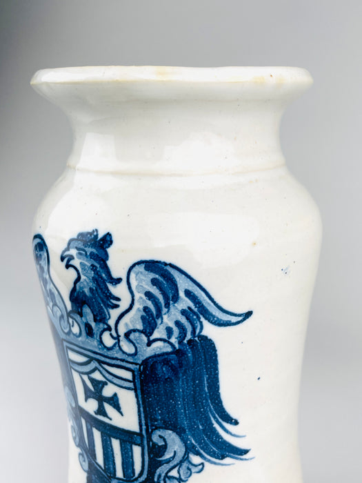 Lower portion of the heraldic decoration on a Spanish blue and white albarello apothecary jar, featuring a shield with a dark cobalt cross and vertical stripes. Curled scroll ornaments frame the shield, while long layered eagle tail feathers extend down the white ceramic body. The hand painted design provides an elaborate graphic contrast to the simple wheel thrown form.