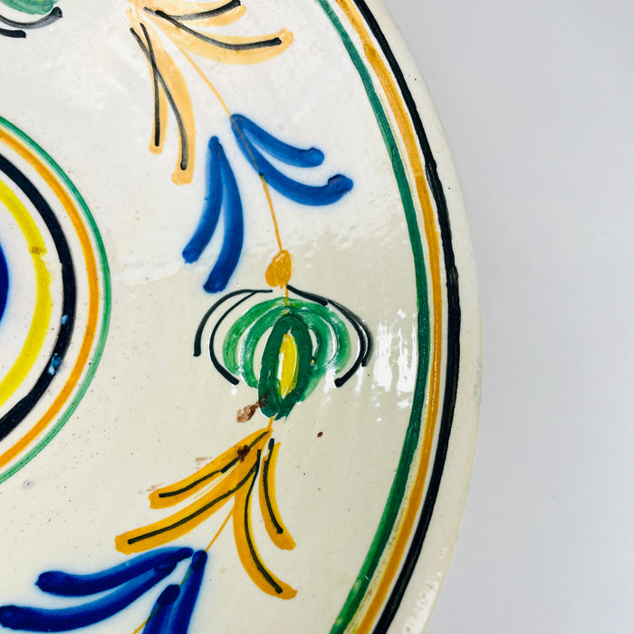 Castilian pottery decoration on an antique lebrillo bowl, with rounded green vegetal motifs edged in black and joined by fine ochre stems. Cobalt blue and honey yellow flourishes fill the spaces between them, while concentric border lines repeat the circular form. The lively palette complements cottage decor while remaining grounded in Spanish folk art.