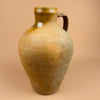 Vintage ceramic jug with a brownish-green glaze on a beige background. Large antique Spanish terracotta cántaro featuring a warm ochre glaze on the upper neck and an unglazed lower body. Ideal for rustic farmhouse or Mediterranean decor, showcasing hand-thrown craftsmanship and organic surface patina.