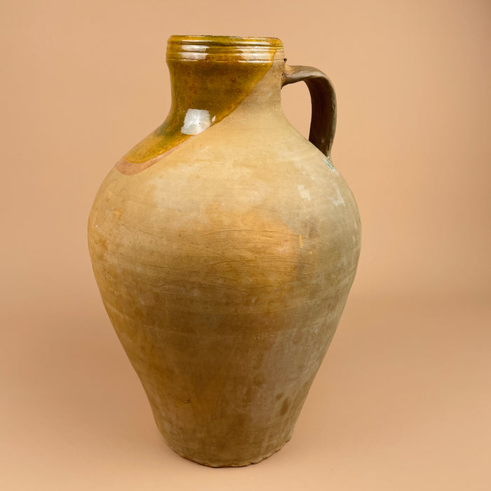 Vintage ceramic jug with a brownish-green glaze on a beige background. Large antique Spanish terracotta cántaro featuring a warm ochre glaze on the upper neck and an unglazed lower body. Ideal for rustic farmhouse or Mediterranean decor, showcasing hand-thrown craftsmanship and organic surface patina.