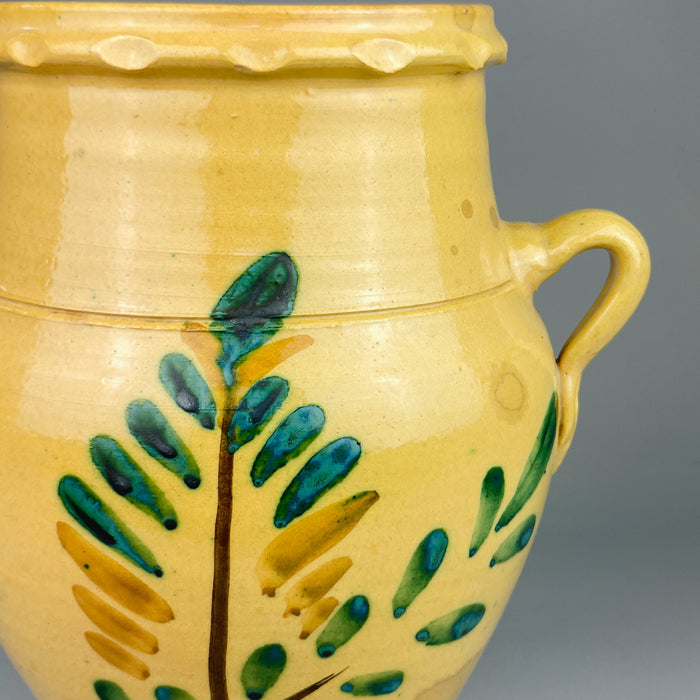 Close-up of the hand-painted foliate motif on the Spanish orza pot, showing green and ochre brushwork applied over the yellow-glazed terracotta body. A rustic Andalusian pottery piece used for storage in traditional Spanish kitchens.