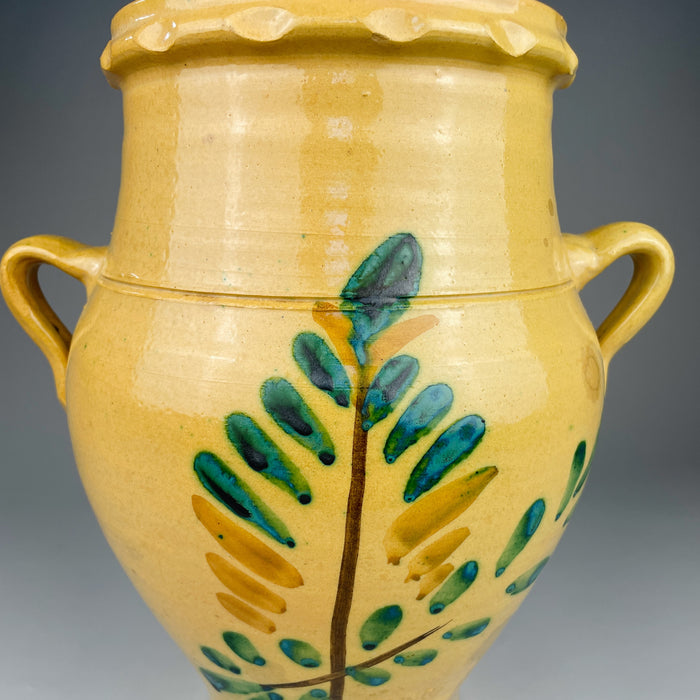 Yellow ceramic vase with green leaf patterns on a gray background. Side angle of the Lucena pottery jar highlighting the curved belly, tapered base, and applied side handles typical of Spanish orzas used for food storage in southern rural homes throughout Andalusia.