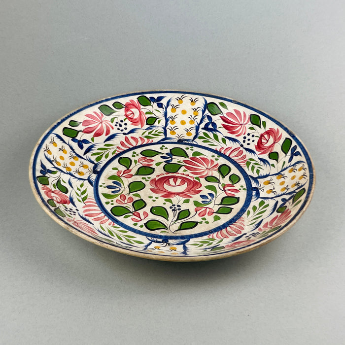 Angled view of antique hand-painted English platter with Arts and Crafts movement-inspired red, green, yellow, and pink floral motifs.