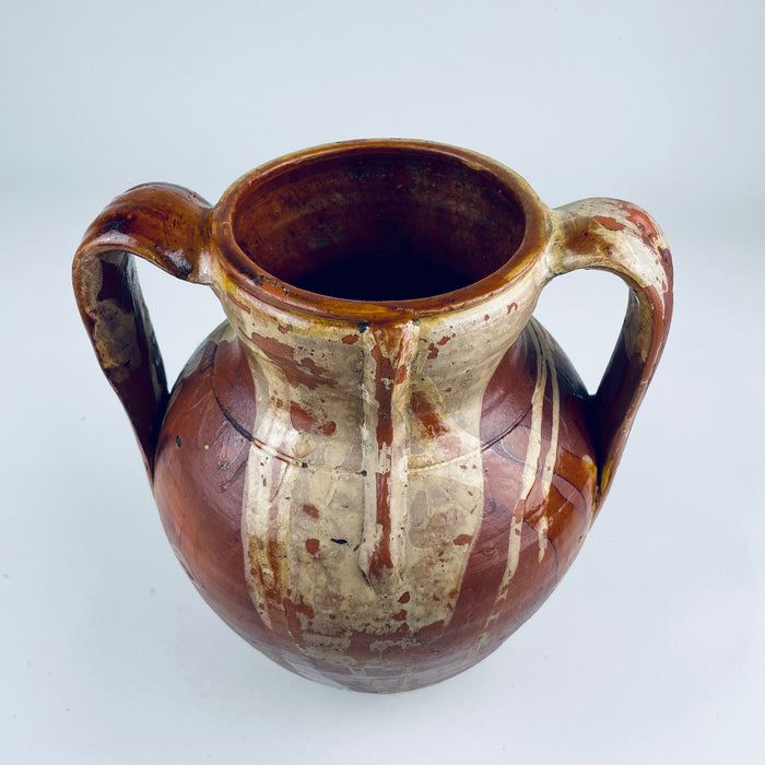 Antique Castilian pottery jar with one of its tall hand applied loop handles visible against the rounded terracotta body. Honey brown glaze covers the shoulder and upper section while the warm red clay remains exposed below, creating the worn two tone character associated with Spanish vernacular pottery and Mediterranean decor.