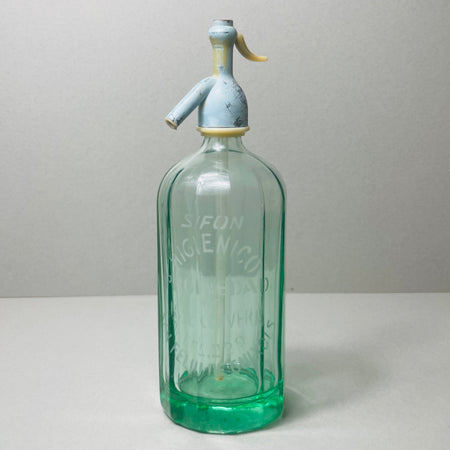 Full view of a vintage Spanish soda siphon made of light green glass, showing elegant etched lettering and original metal siphon top with aged blue patina, photographed on a light background.