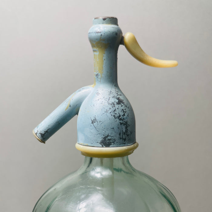 Unique collector’s piece with visible embossed script and aged coloration on top, appealing to lovers of vintage European glass. the bottle’s classic soda siphon shape and retro details like the ivory siphon lever and painted metal top.