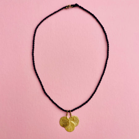 Angled close-up highlighting the hammered coin pendants, each 2 cm wide, reflecting light with a golden shimmer. The artisanal surface texture contrasts with the matte black braided cord, underscoring its role as lightweight minimalist jewelry.