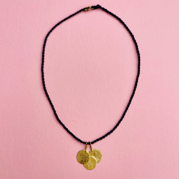 Angled close-up highlighting the hammered coin pendants, each 2 cm wide, reflecting light with a golden shimmer. The artisanal surface texture contrasts with the matte black braided cord, underscoring its role as lightweight minimalist jewelry.