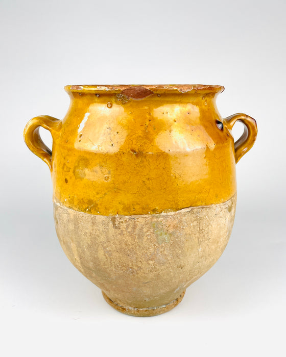 Antique French terracotta confit pot dating to the 19th century, with a rounded body, two curved loop handles and a rich mustard yellow glaze covering the upper half. The lower section retains exposed sandy beige earthenware with extensive surface patina, and the vessel stands on a small circular foot. A traditional French pottery jar with strong sculptural character, suited to French country decor.