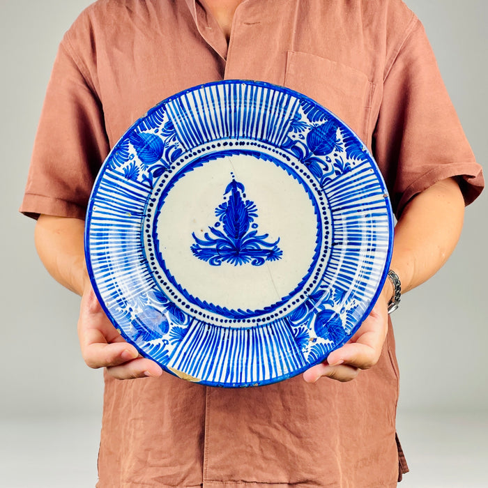 Antique Spanish faience plate attributed to Manises, Valencia, with cobalt blue painting on an opaque warm white tin glaze. The deep charger displays handmade variation in line spacing, pigment density and floral brushwork, together with rim chips, glaze losses, crazing and a historic repaired break that should remain part of its presentation.