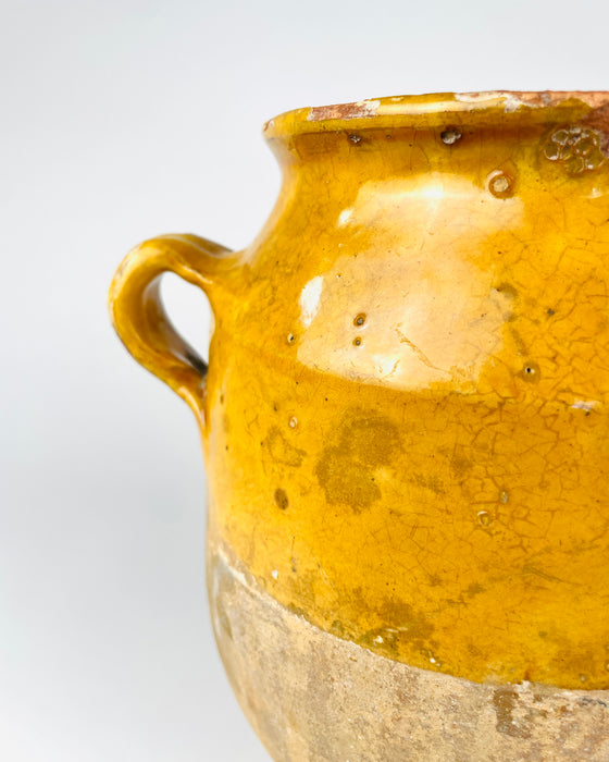 Applied handle of an antique French yellow glazed pottery jar showing the thick curved terracotta loop, worn edges and small losses revealing the clay beneath the glaze. The surrounding mustard coloured surface contains firing marks, minute pits and fine crackling, characteristic details of this weathered confit vessel. An original utilitarian French ceramic object for farmhouse decor.