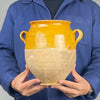 Detail of a 19th century French confit pot showing the curved terracotta handle, flared rim and golden yellow glazed shoulder. Fine glaze crazing, small firing bubbles and worn areas reveal the handcrafted character of this antique earthenware vessel. The warm ochre surface contrasts with the pale unglazed clay below, making this traditional French storage jar suitable for farmhouse decor.