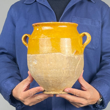 Detail of a 19th century French confit pot showing the curved terracotta handle, flared rim and golden yellow glazed shoulder. Fine glaze crazing, small firing bubbles and worn areas reveal the handcrafted character of this antique earthenware vessel. The warm ochre surface contrasts with the pale unglazed clay below, making this traditional French storage jar suitable for farmhouse decor.