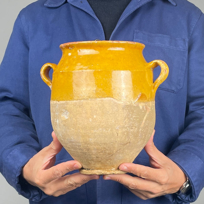 Detail of a 19th century French confit pot showing the curved terracotta handle, flared rim and golden yellow glazed shoulder. Fine glaze crazing, small firing bubbles and worn areas reveal the handcrafted character of this antique earthenware vessel. The warm ochre surface contrasts with the pale unglazed clay below, making this traditional French storage jar suitable for farmhouse decor.