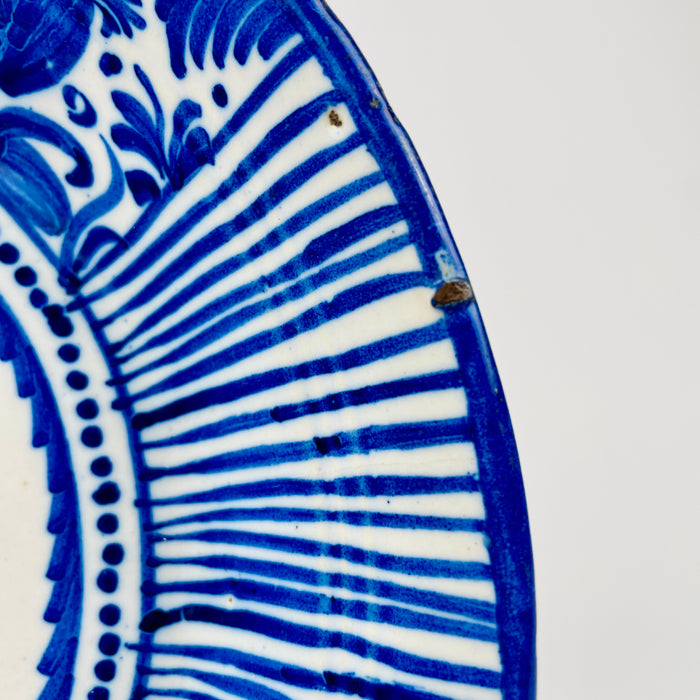 Cobalt blue Manises plate from 19th-century Spain, made in tin-glazed earthenware and painted with a central stylized botanical motif surrounded by dotted bands, fruit forms and tightly spaced radial stripes. Its saturated blue and cream palette makes the antique ceramic effective as Mediterranean wall art or Spanish pottery decor.