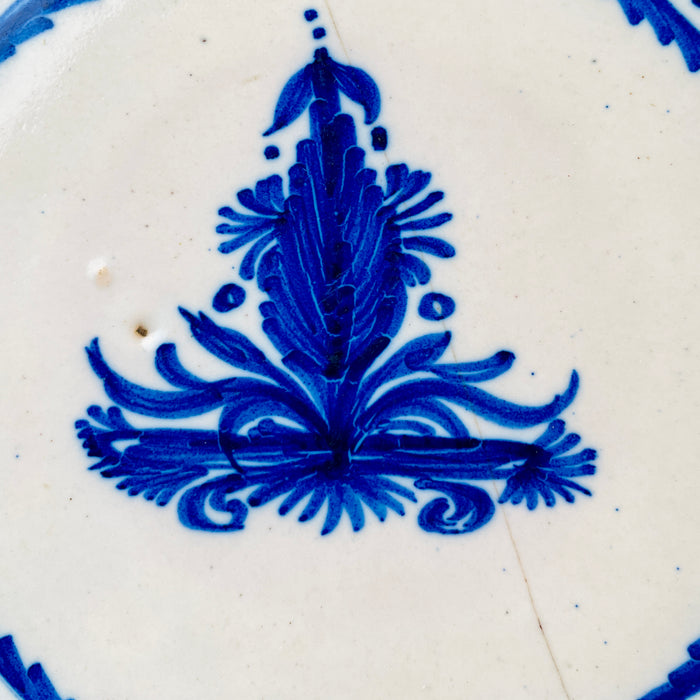 Nineteenth-century Spanish Manises charger in blue and white majolica, hand painted with a stylized central palmette, scrolling foliage, fruit maotifs, dotted borders and alternating fields of cobalt radial lines. The decorative plate combines strong Mediterranean colour with visible rim wear and a preserved historic staple repair for characterful ceramic wall art.
