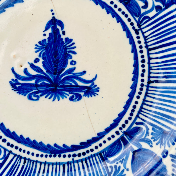 Central decoration on a 19th-century Spanish Manises plate, hand painted in deep cobalt blue over opaque white tin glaze. The symmetrical foliate motif resembles a stylized palmette or flower rising above scrolling leaves and small rosettes, forming the focal point of this antique majolica charger and Mediterranean ceramic wall art.