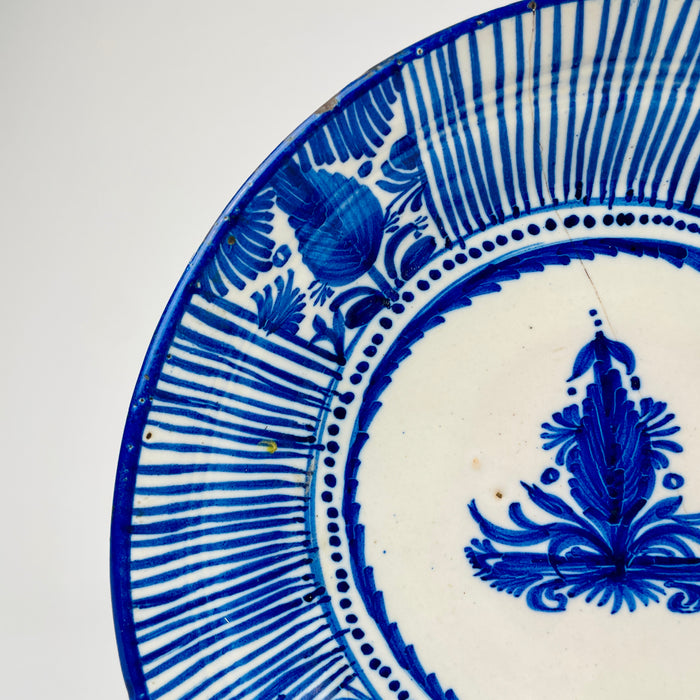 Antique Spanish majolica plate with a five-centimetre depth, slightly raised foot and gently curved earthenware body. The cobalt blue rim and hand-painted Manises decoration continue around the interior, while the reverse carries an F.M.-style mark and historic metal staple repair associated with the plate’s long working life.