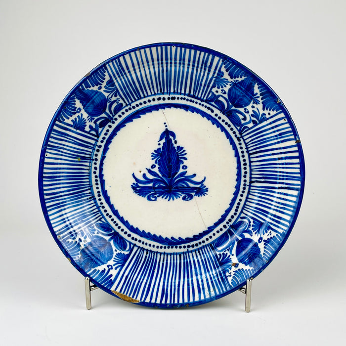 Antique Manises pottery plate from Spain with cobalt blue brushwork on a creamy tin-glazed ground. The broad decorative charger features alternating bands of fine radial stripes and stylized botanical motifs around a central floral medallion, while age-related rim wear and glaze losses preserve the character of this 19th-century Spanish ceramic wall plate.
