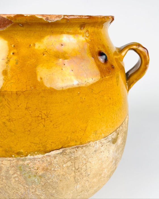 Applied handle of an antique French yellow glazed pottery jar showing the thick curved terracotta loop, worn edges and small losses revealing the clay beneath the glaze. The surrounding mustard coloured surface contains firing marks, minute pits and fine crackling, characteristic details of this weathered confit vessel. An original utilitarian French ceramic object for farmhouse decor.
