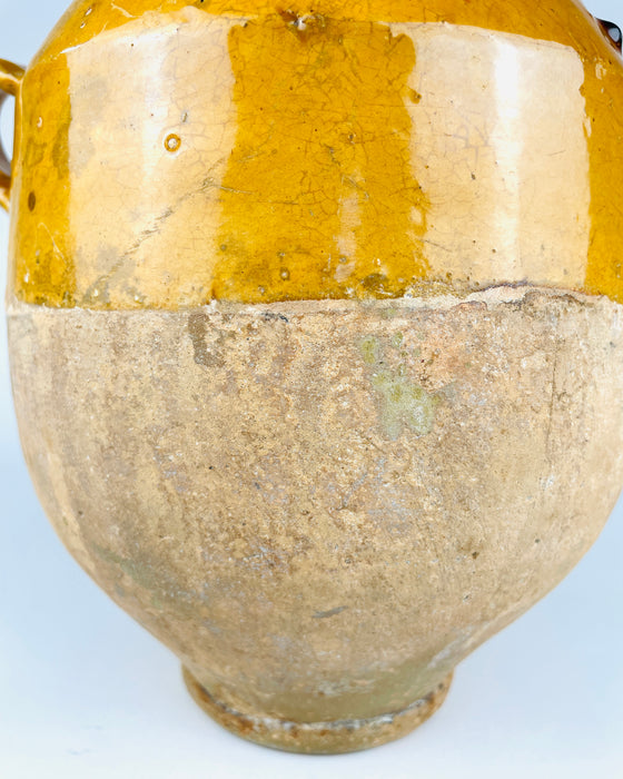 Antique French confit pot with a rounded body and an applied terracotta loop handle showing patches of worn golden glaze and exposed clay. The mustard yellow upper surface has fine crazing, small firing imperfections and the natural tonal variation of traditional pottery. The partially unglazed lower body reflects the practical construction of an old French preservation jar for French provincial decor.