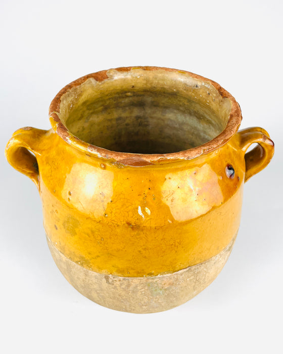 Lower section of a 19th century French terracotta confit jar showing the softly rounded body, rough beige earthenware surface and small raised circular foot. The upper edge of the natural clay meets an irregular band of mustard yellow glaze, highlighting the partially glazed construction of traditional French preservation pots. A characterful antique ceramic vessel for rustic home decor.
