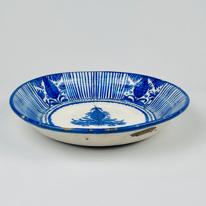 Antique Spanish ceramic charger attributed to Manises, with a deep rounded profile and hand-painted cobalt blue decoration across a warm cream tin-glazed surface. Alternating striped sections and stylized floral or fruit motifs frame the central palmette, while the repaired crack, rim fritting and glaze losses document extensive age and use.