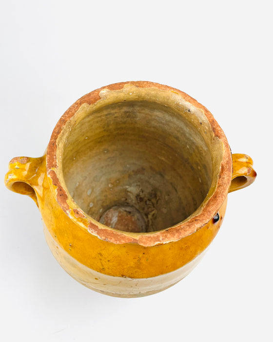 A substantial antique French terracotta confit pot held with two hands to demonstrate its scale, revealing the warm mustard yellow glazed upper body, natural beige clay lower section and two symmetrical handles. The rounded vessel has a flared lip and narrow circular foot, with surface patina throughout. This traditional 19th century French pottery jar is well suited to French country decor.