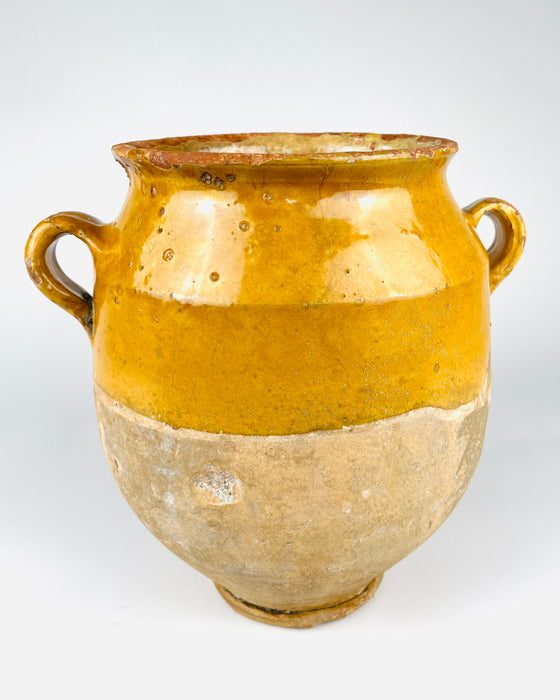 Surface detail of an antique French confit pot showing deep golden ochre glaze, fine interconnected crazing and small irregularities in the ceramic finish. Below the glazed section, the natural terracotta has developed a pale textured patina with areas of mineral staining and abrasion. The contrast between glossy colour and weathered clay makes this 19th century vessel suitable for wabi sabi decor.