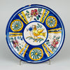 Large antique Spanish majolica wall plate from Manises, Valencia, displayed above an iron bed as ceramic wall art. The 31.5 cm tin glazed earthenware charger features a stylized golden yellow bird framed by cobalt blue bands, pink flowers, green leaves and yellow radial panels, creating vivid Mediterranean decor.