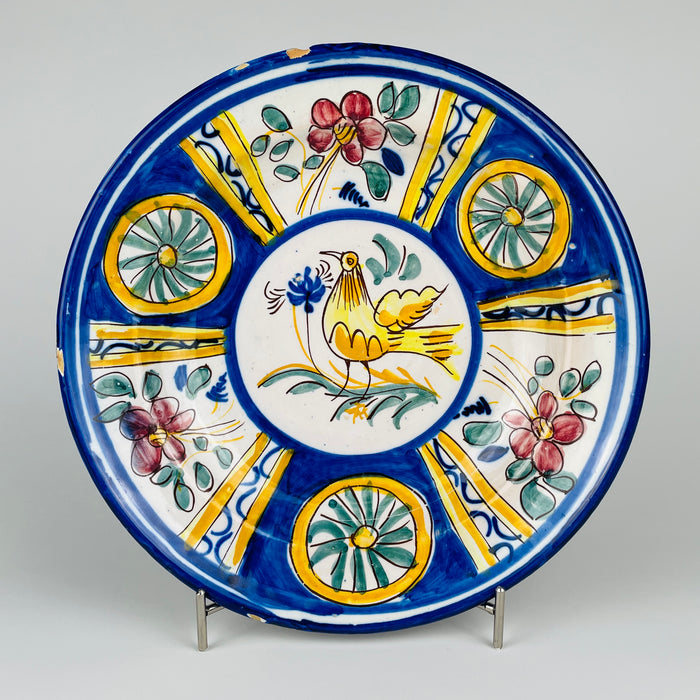 Large antique Spanish majolica wall plate from Manises, Valencia, displayed above an iron bed as ceramic wall art. The 31.5 cm tin glazed earthenware charger features a stylized golden yellow bird framed by cobalt blue bands, pink flowers, green leaves and yellow radial panels, creating vivid Mediterranean decor.