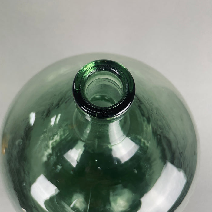 Light reflecting off the handblown green glass of the demijohn, showing authentic farmhouse design from mid-20th century France. French vintage glass wine vessel with tall, slim bottleneck and wide body, ideal for collectors or rustic interior décor.