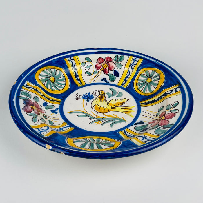 Central motif of the antique Manises wall plate, showing a stylized golden yellow bird with a long tail, fine brown feather lines, slim legs and a cobalt blue flower near its beak. Green leaves and sweeping stems complete the hand painted scene on white tin glazed earthenware from Valencia, Spain.