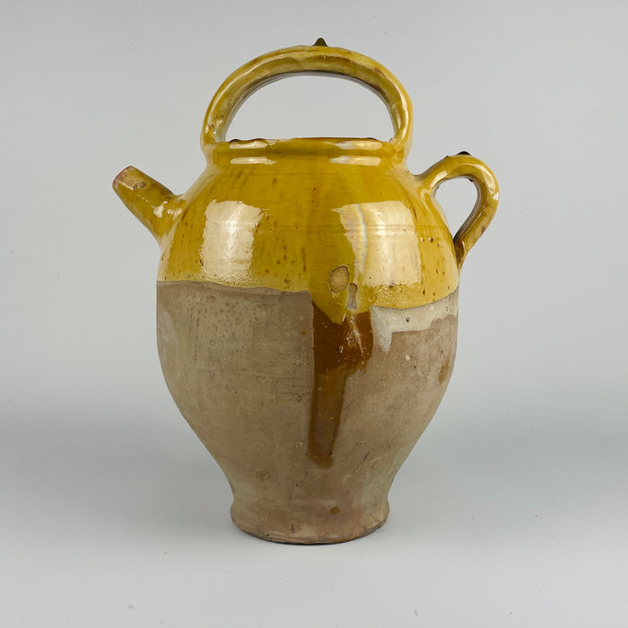 Side profile of a large antique French terracotta cruche pot shows its curved shoulder, yellow glazed surface, and spout used historically for pouring water or wine. The pot’s aged surface displays natural wear consistent with 19th century use. This handcrafted French pottery piece enhances farmhouse decor, cottage decor, rustic home decor, french country decor, and mediterranean decor, especially in rooms where warm terracotta tones are desired.