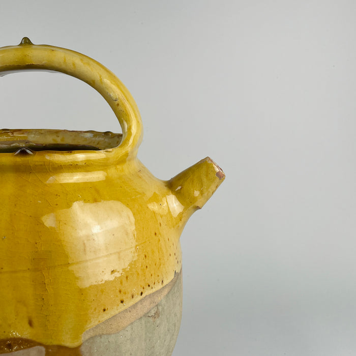 This angle captures the sculptural silhouette of a yellow glazed terracotta cruche, a classic form in antique farmhouse decor and French country decor. The glaze shows age patina, with minor imperfections typical of utilitarian pottery from the 1800s, offering strong visual presence for rustic home decor enthusiasts.