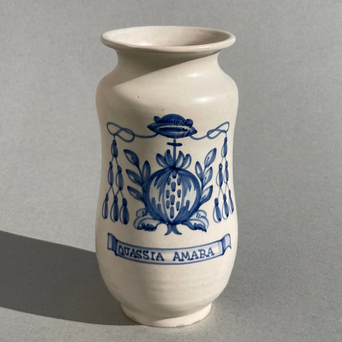 Spanish Talavera apothecary jar, ceramic vessel with blue and white glaze, frontal view showing hand-painted pomegranate motif and ribbon with “Quassia Amara,” ideal for vintage pharmacy decor or herbal collections.