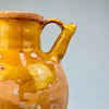 Close-up of a ceramic pitcher with a yellowish-brown glaze on a gray background. Angled view of antique French gargolette jug with rich ochre glaze, rustic patina, and exposed terracotta surface, styled for use as a sculptural accent or as a decorative water vessel in farmhouse-inspired settings.