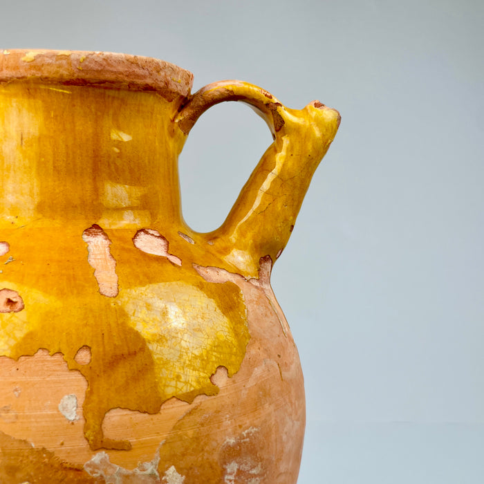 Close-up of a ceramic pitcher with a yellowish-brown glaze on a gray background. Angled view of antique French gargolette jug with rich ochre glaze, rustic patina, and exposed terracotta surface, styled for use as a sculptural accent or as a decorative water vessel in farmhouse-inspired settings.